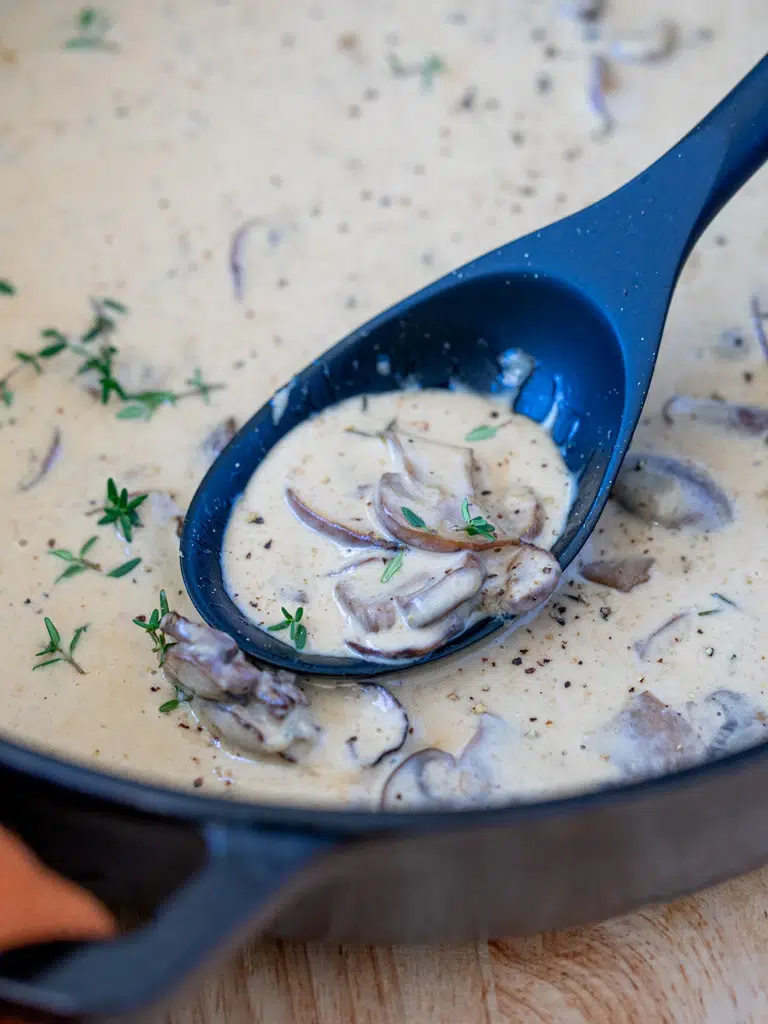 Close-up of creamy mushroom sauce with sliced mushrooms and fresh thyme in a black ladle over a skillet, ready for serving.