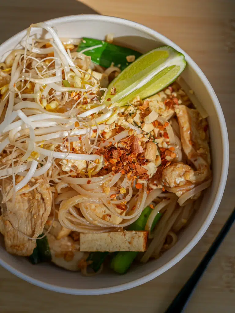 A Delicious Bowl of chicken Pad Thai garnished with chopped peanuts and lime wedges.