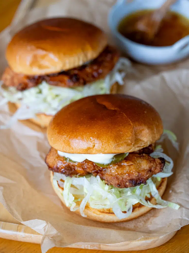 Two hot honey crunch chicken burgers with crispy fried chicken, shredded lettuce, pickles, and mayo on toasted brioche buns, served on parchment paper.