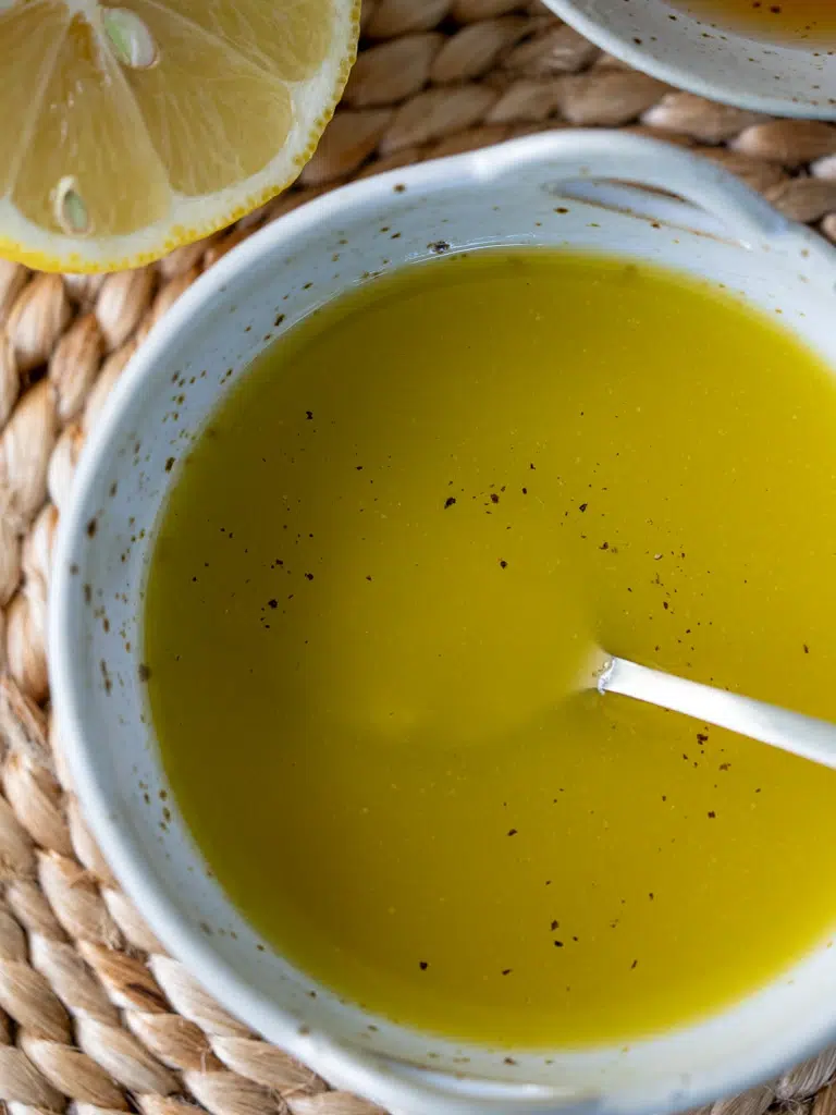 Close-up of a simple lemon vinaigrette with black pepper in a white ceramic bowl, next to a halved lemon.